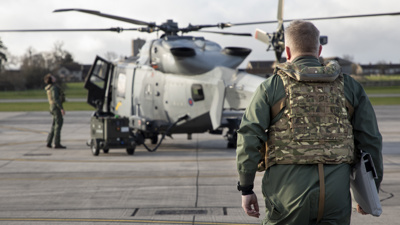 A helicopter pilot in a military green flight suit, wearing a multi-terrain pattern emergency parachute system, is holding a grey document bag. He is walking towards a helicopter, which another soldier is standing next to.