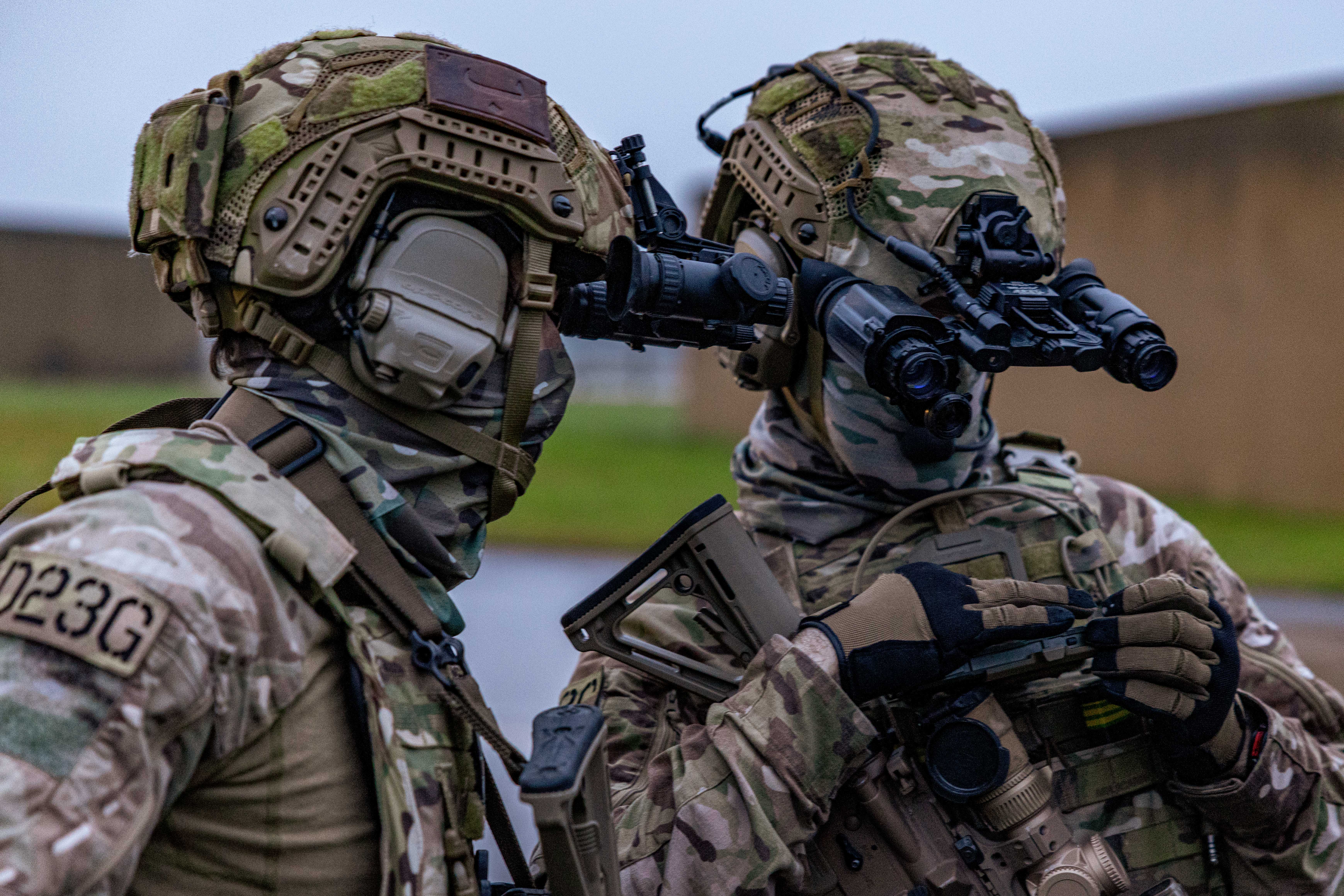 Two soldiers in full camouflage gear and helmets communicate closely during a tactical operation outdoors.