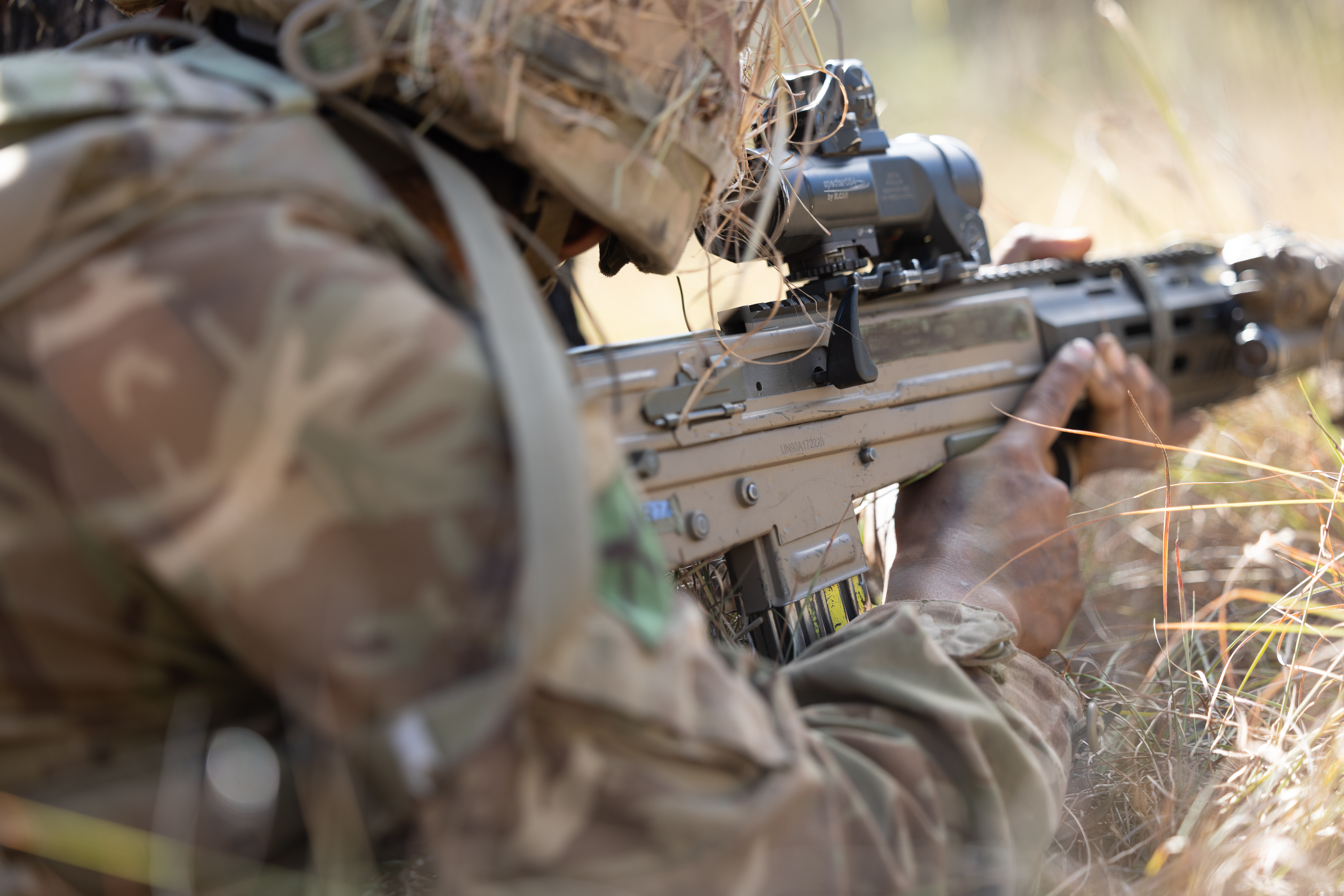  Soldier in camouflage gear aiming a scoped rifle while lying prone in tall grass during daylight.