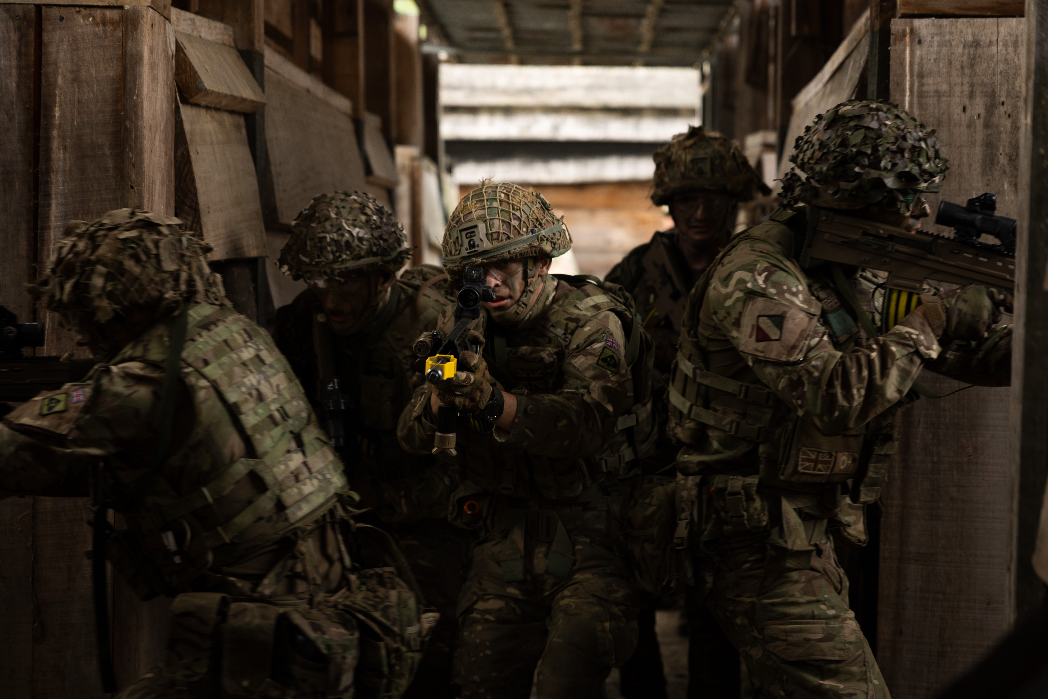 Group of soldiers in camouflage gear tactically advancing through a narrow, dimly lit corridor during a training exercise.