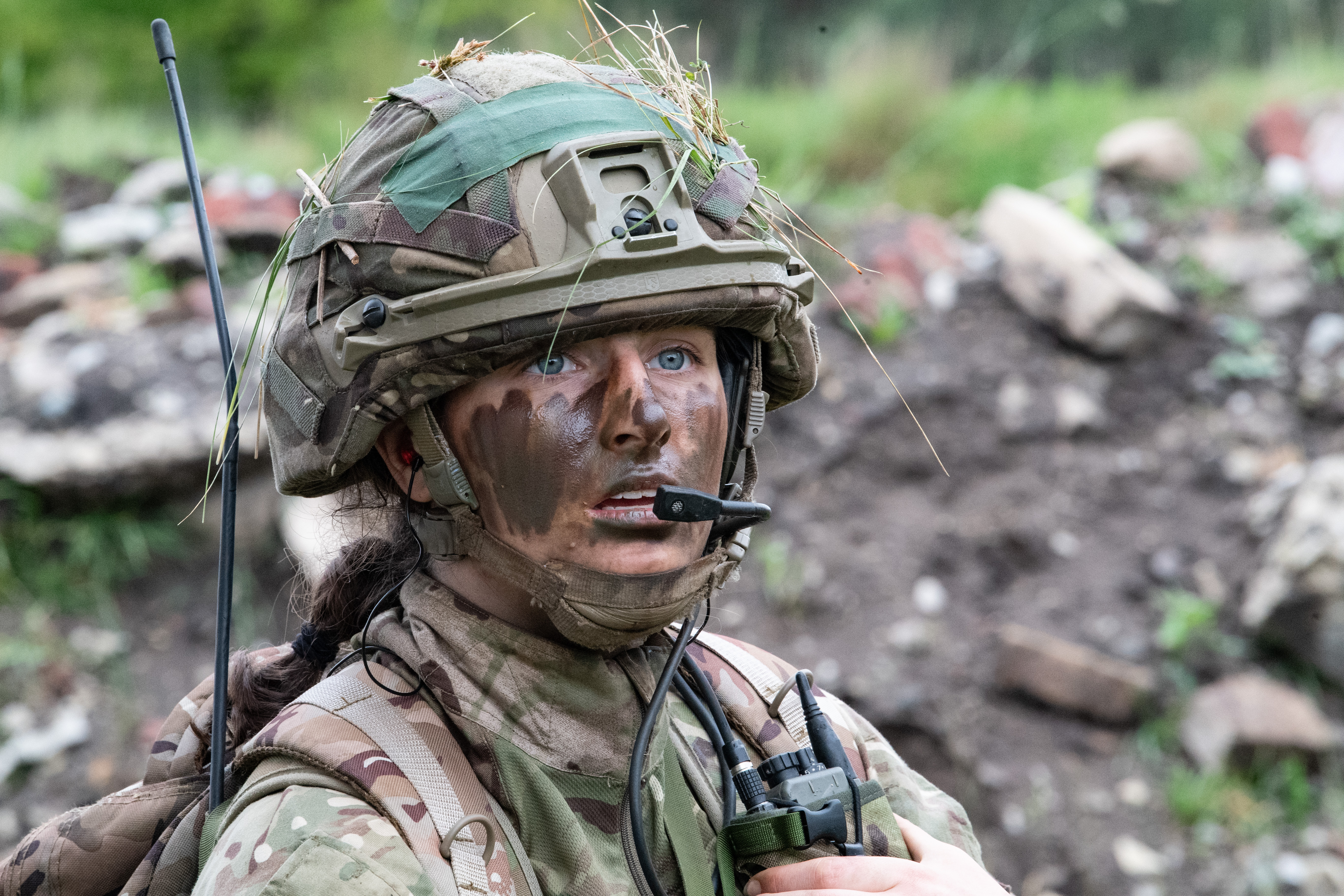 Soldier in camouflage gear and helmet with radio equipment in an outdoor environment.